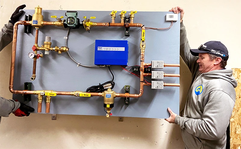 Two installers lifting a pre-piped mechanical board into position on a stud wall during a radiant heating installation