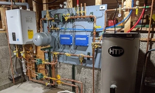 A finished radiant heating mechanical room: wall hung boiler, expansion tank, circulators and controls on a mounted board, and an indirect water heater